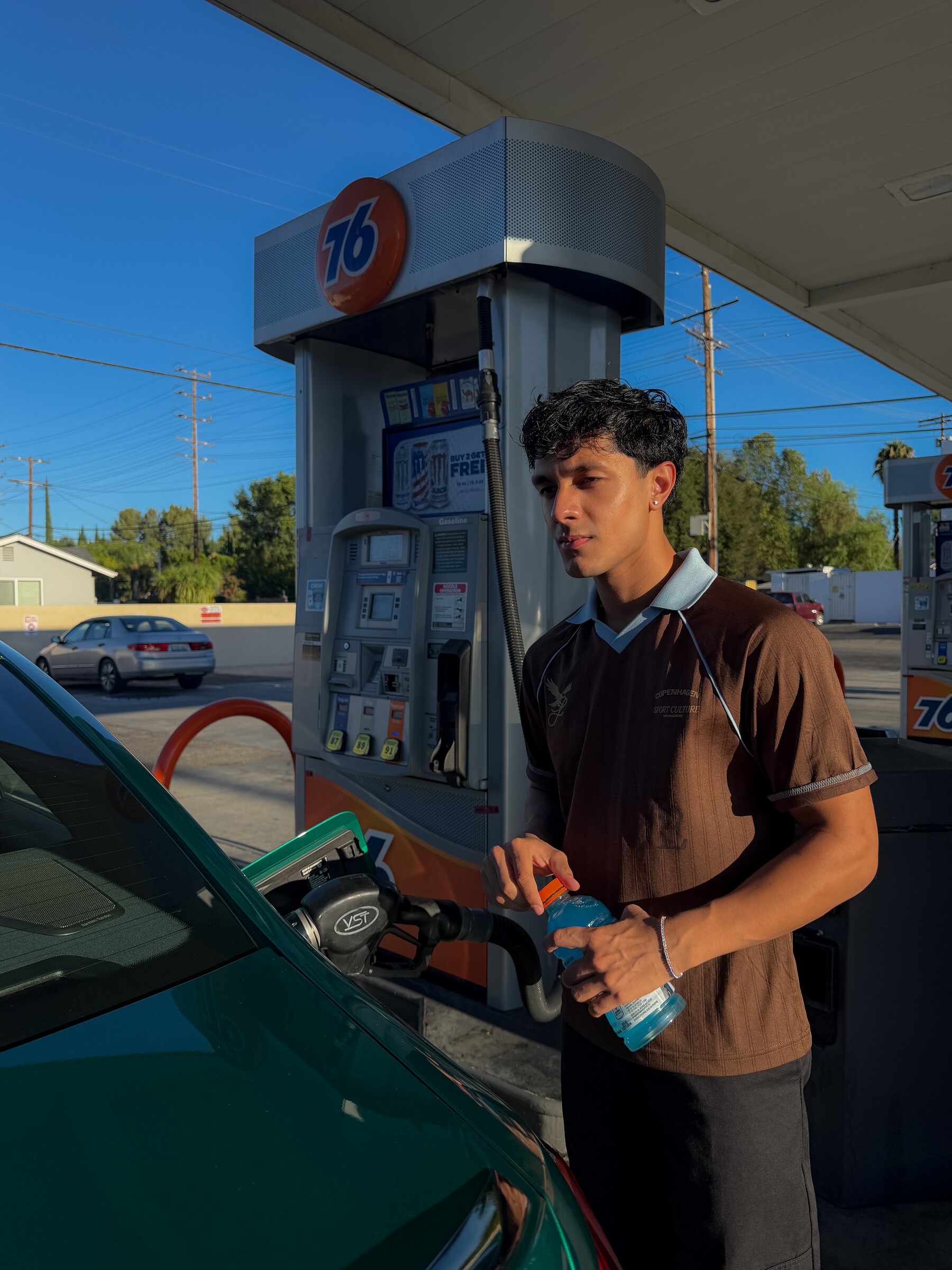 Aryan Mahajan in a brown jersey beside a green BMW M3 at a gas station