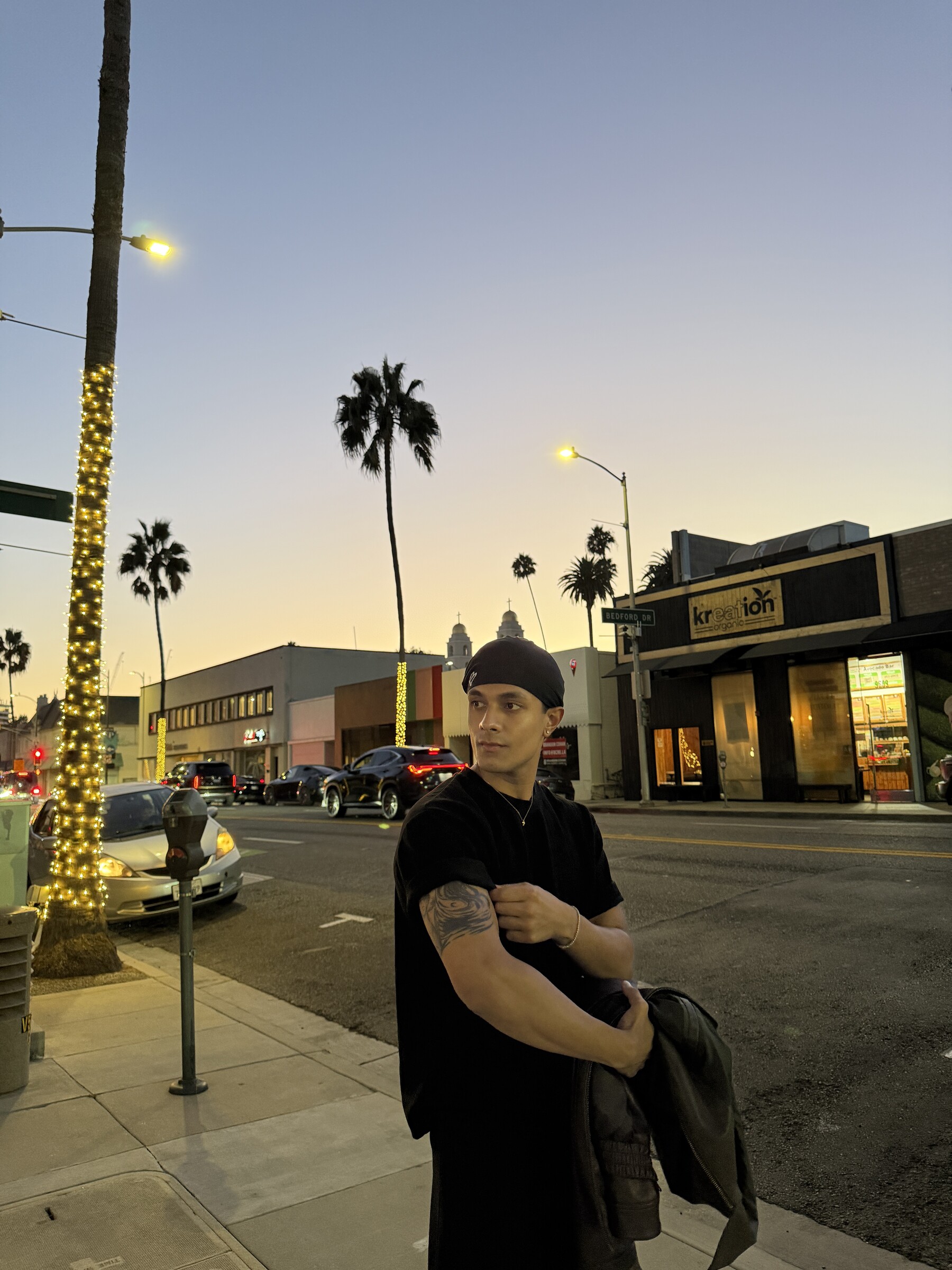 Aryan Mahajan wearing black and carrying a jacket on a palm-lined street